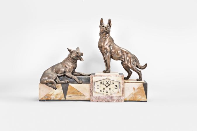 Clocks- 2 (Two) French: (1) France, for Brunott Juwelier, Rotterdam, Netherlands, three piece mantel garniture, 8 day, time and strike, spring driven pendule de Paris movement in an Art Deco case made of white onyx and gray marble, topped with the patinated figure of a woman with outstretched arms flanked by seated musicians on either side, gold painted metal dial, stylized arabic numerals and black steel hands, with two matching side pieces, c1920; (2) France, figural mantel clock, 8 day, time and strike, spring driven pendule de Paris movement in an Art Deco case made of white onyx, black slate, and variegated roseate marble, topped with two large cast German Shepard dog figures, silver colored metal dial with stylized arabic numerals and black steel hands, c1925.