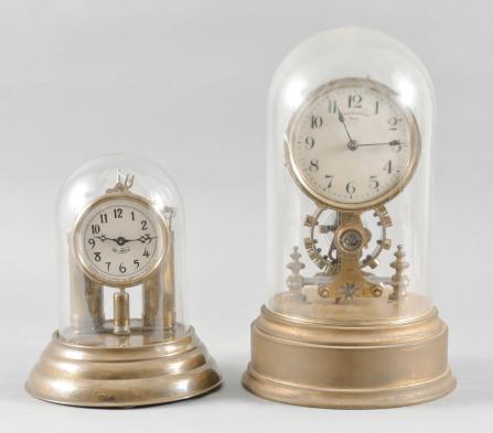 Clocks- 2 (Two) Electromechanical: (1) Mountain State Electrical Co., Wheeling, W.V., "No-Key" mantel timepiece, weight driven clock with battery-powered electromagnet for periodic rewinding, molded brass base, celluloid dial, and blued steel hands, all under a glass dome, c1910; (2) Eureka Clock Co. Ltd., London, England, electromechanical timepiece, long-running, battery powered tall movement with large exposed balance oscillating below the arabic numeral white enamel dial with blued steel hands, all resting on a hollow brass base, and covered with a glass dome, c1906.