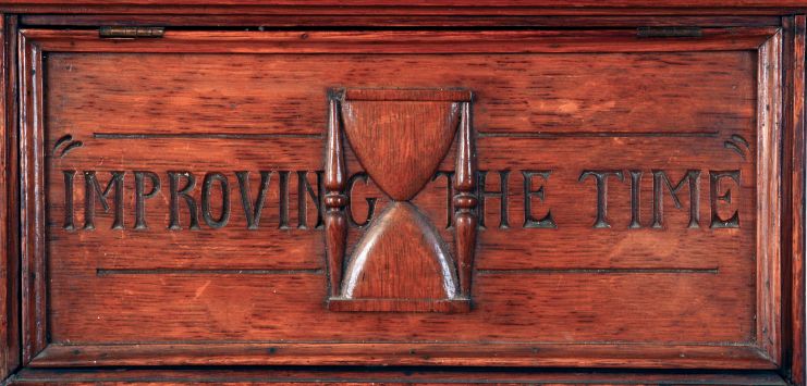 Folk Art wall clock, large, well made oak case with gabled facade, incised line carving, ogee and astragal moldings, and hinged access panel for pendulum with applied, carved hourglass, and also carved "Improving The Time", top with brass finial, Roman numeral paper dial, spade and poker hands, 8 days spring driven, timepiece movement by Ansonia, case dated in pencil on back, 1882