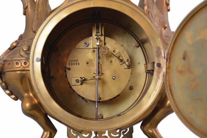 France, figural brass mantel clock, 8 day, time and strike on a pierced base with two short pillars supporting winged griffon figures in profile flanking a central cylinder holding an ornate brass dial with blued steel hands and white enameled cartouche numerals in blue, surmounted by a decorative urn. The pendule de Paris movement with partially exposed pendulum is stamped "CHLES MT".